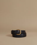 A black leather belt with a polished gold-tone metal buckle, neatly coiled on a warm beige studio background.