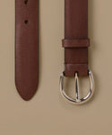 A close-up of a brown leather belt showing the strap end and buckle against a solid beige background.