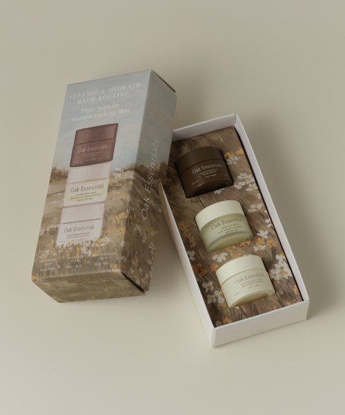A boxed set of three Oak Essentials skincare products displayed on a warm beige background. The set includes a tall outer box with a floral field illustration and a white inner tray holding two cream-colored jars and one dark brown jar, all labeled Oak Essentials.