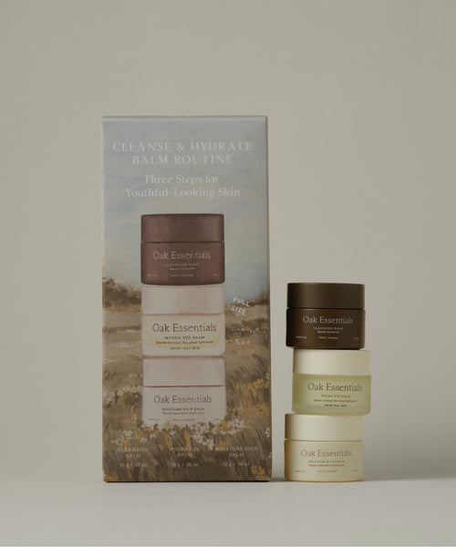 Oak Essentials Cleanse & Hydrate Balm Routine set, featuring a tall illustrated outer box and three stacked cosmetic jars in dark brown, cream, and light beige tones against a solid taupe background.