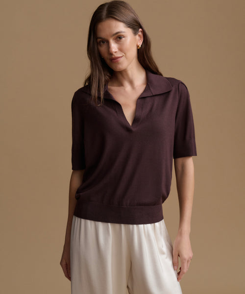 A woman modeling a short-sleeve knit polo top in deep plum purple, paired with cream-colored pants, against a solid warm beige studio background.