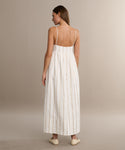 A woman with straight light brown hair wears the Cove Dress, a white casual dress with vertical gold stripes and thin straps, paired with light-colored flats. She faces away against a neutral background.