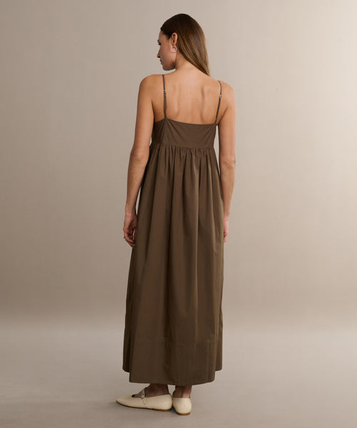 A woman with straight brown hair stands facing away, wearing the Cove Dress—a relaxed fit, brown cotton dress with thin straps—and cream-colored flat shoes against a plain beige background.