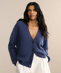 A woman with long dark hair wears the Cropped Cashmere Cocoon Cardigan in deep blue and white pants, standing against a beige background. She gazes slightly to the side with a relaxed expression.