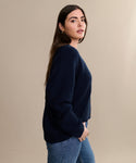 A woman with long brown hair models the Cropped Cashmere Cocoon Cardigan in navy with blue jeans, standing sideways with one hand in her pocket against a plain beige background.