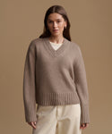 A woman wearing a taupe brown ribbed knit V-neck sweater over a cream undershirt and cream trousers, standing against a solid tan studio background.