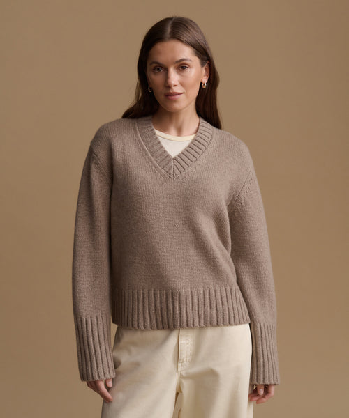 A woman wearing a taupe brown ribbed knit V-neck sweater over a cream undershirt and cream trousers, standing against a solid tan studio background.