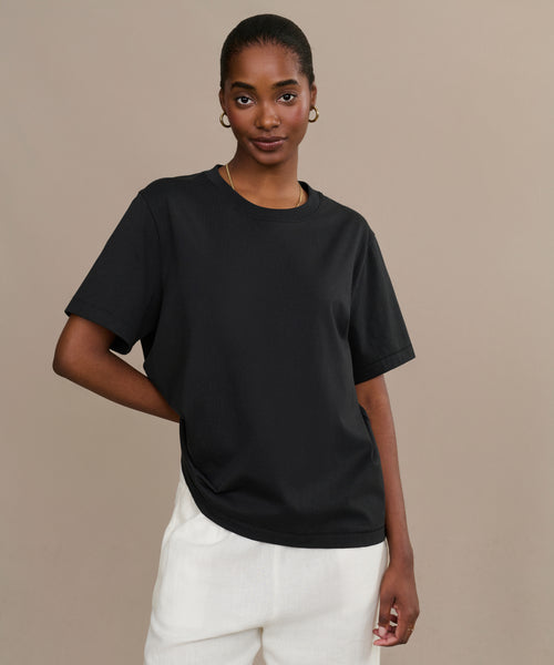 A woman stands against a plain beige background, wearing the Crosby Tee and white pants. Made from premium jersey cotton, the relaxed fit tee complements her confident stance as she poses with her left hand on her hip.