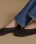 A close-up of a person wearing dark brown suede moccasin-style flats with blue jeans, set against a solid tan background.