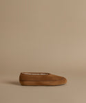 A single tan suede moccasin slipper with contrast stitching and a white lining, shown in profile against a solid beige background.