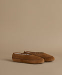A pair of tan suede slip-on slippers with whipstitching and a soft white lining, displayed on a solid beige background.