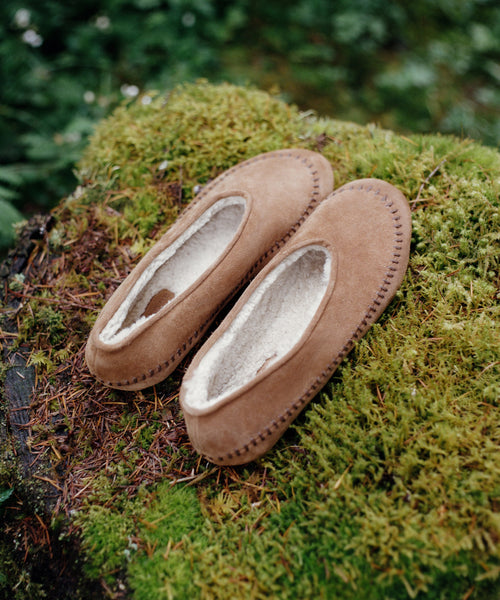 A pair of tan suede moccasin-style slippers with white shearling lining, resting on a mossy log outdoors.