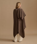 A woman shown from the back wearing a long, draped brown knit poncho or cape over cream wide-leg pants, standing against a plain beige studio background.