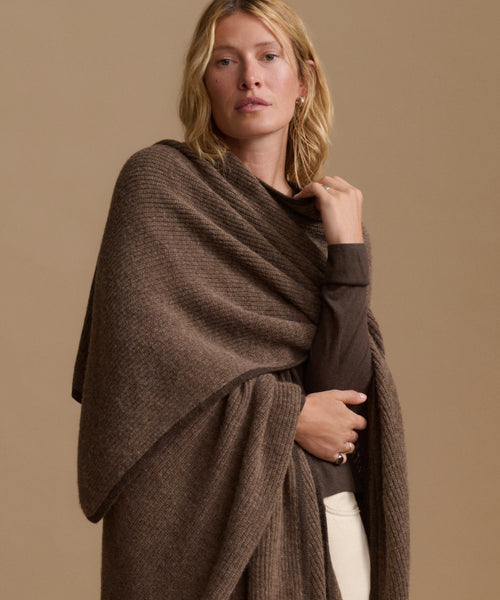 A woman modeling a chunky knit brown shawl wrap draped over her shoulders, paired with a dark brown long-sleeve top and cream pants, against a solid beige studio background.