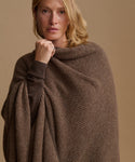 A woman wearing a brown ribbed knit wrap or poncho over a dark brown top, photographed against a plain warm beige studio background.