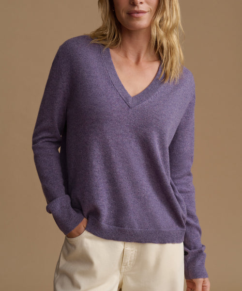 A woman wearing a heathered purple V-neck knit sweater and cream pants, standing against a solid tan background.