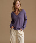 A woman wearing a heathered purple long-sleeve V-neck sweater and cream-colored pants, standing against a solid tan studio background.