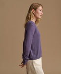 A woman in side profile wearing a heathered purple knit sweater and cream trousers, set against a solid warm beige studio background.