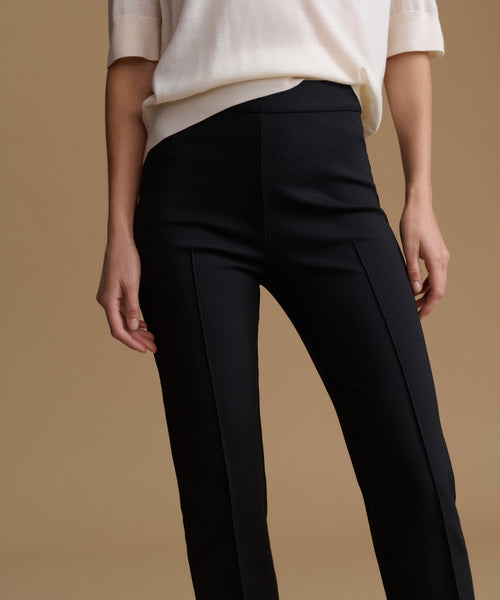 A model wearing black high-waisted trousers with front pleats and a cream-colored short-sleeve top, set against a solid warm beige background.