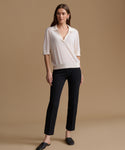 A woman stands against a warm beige studio background wearing a cream short-sleeve knit polo-style top and black cropped trousers with dark loafers.