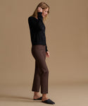 A woman stands in profile wearing a black long-sleeve button-front top, brown cropped pants, and black slip-on shoes against a plain warm beige studio background.