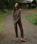 A woman stands on a gravel path in a wooded outdoor setting, wearing a ribbed brown mock-neck sweater, brown pants, and brown loafers.
