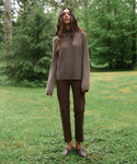 A woman stands outdoors in a grassy park area with trees in the background, wearing a ribbed brown turtleneck sweater, dark brown pants, and brown loafers.