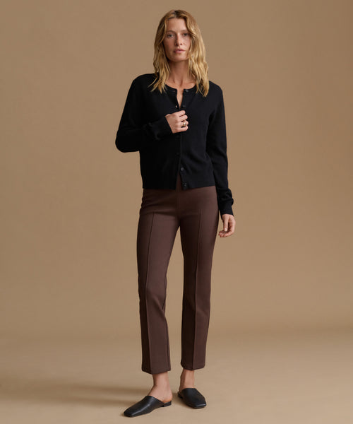A woman modeling a black button-up cardigan and brown cropped trousers with black mule flats, set against a solid beige studio background.