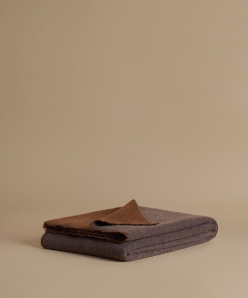 A folded dark gray wool blanket with a contrasting brown color on top, displayed on a warm beige studio background.
