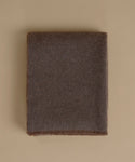 A folded dark brown woolen blanket with fringed edges, displayed flat against a solid beige background.