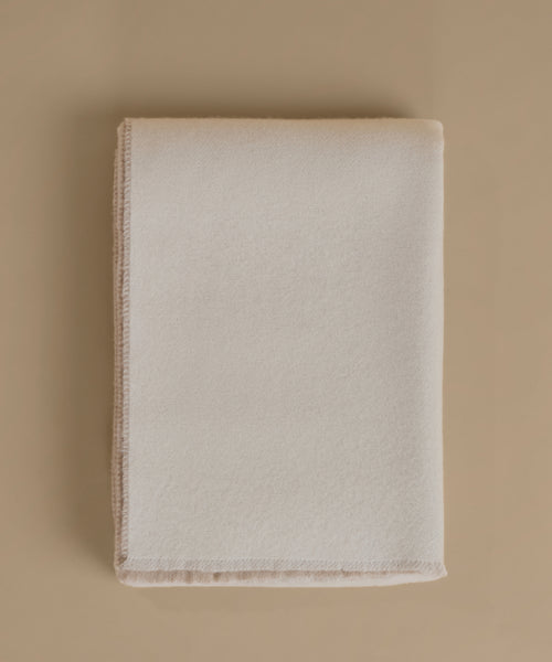 A folded cream-colored wool blanket with a soft, brushed texture and fringed edges, displayed on a solid beige background.