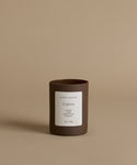 A glass candle in th scent Cypress in a brown vessel against a beige background.