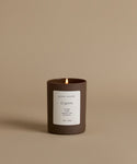 A lit candle in a matte dark brown glass jar with a white label reading 'jenni kayne' and 'Cypress', set against a solid warm beige background.