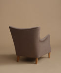 A single upholstered armchair shown from the back, upholstered in a textured taupe fabric with light natural wood legs, set against a solid warm beige studio background.