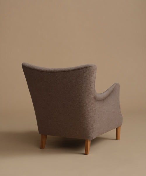 A single upholstered armchair shown from the back, upholstered in a textured taupe fabric with light natural wood legs, set against a solid warm beige studio background.