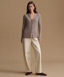 A woman stands against a warm beige studio background wearing a taupe ribbed button-front cardigan, cream wide-leg pants, and dark brown slip-on shoes.