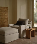A Harbor Chair in Drift Linen with a matching ottoman sits beside a wooden side table topped with a vase of white flowers, in a softly lit room featuring wood-paneled walls and a large window.