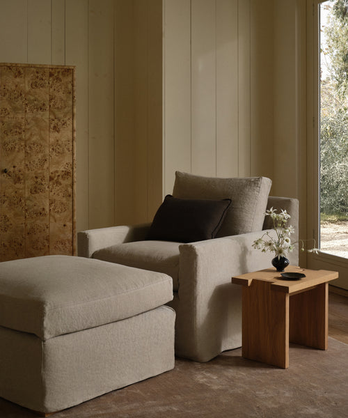 A Harbor Chair in Drift Linen with a matching ottoman sits beside a wooden side table topped with a vase of white flowers, in a softly lit room featuring wood-paneled walls and a large window.