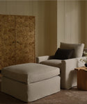 A cozy living space with a light beige armchair, Harbor Ottoman by Jenni Kayne Furniture, dark accent pillow, wooden side table with a vase of flowers, and a decorative wood-paneled screen against cream-colored walls.