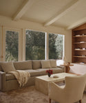 A cozy living room featuring the Harbor Sofa, handmade in Los Angeles with linen upholstery, a cream armchair, marble coffee table topped with flowers, built-in wood shelves, and large windows overlooking a lush garden.