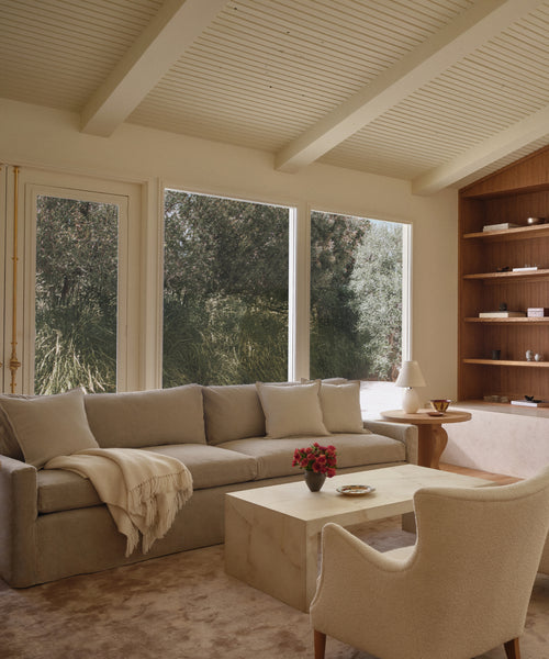 A cozy living room featuring the Harbor Sofa, handmade in Los Angeles with linen upholstery, a cream armchair, marble coffee table topped with flowers, built-in wood shelves, and large windows overlooking a lush garden.