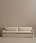 The Harbor Sofa, handcrafted in Los Angeles with beige linen fabric, includes two seat and two back cushions, creating a minimalist vibe against a plain taupe wall.