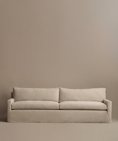 The Harbor Sofa, handcrafted in Los Angeles with beige linen fabric, includes two seat and two back cushions, creating a minimalist vibe against a plain taupe wall.