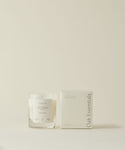 The Santo Wood Candle, featuring its signature glass design and woody scent, is displayed next to a white box labeled "Oak Essentials," all set against a simple light beige background.