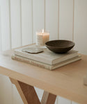 A light wooden table holds two stacked books, a smartphone, a burning Santo Wood Candle in a glass jar, and a dark ceramic bowl, all neatly arranged against a backdrop of white vertical paneling.