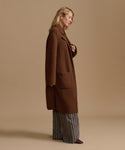 A woman stands in profile wearing a long brown wool-blend coat over wide-leg striped trousers and dark shoes, photographed against a plain warm beige studio background.