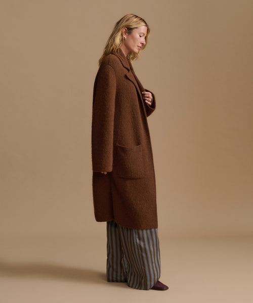 A woman stands in profile wearing a long brown wool-blend coat over wide-leg striped trousers and dark shoes, photographed against a plain warm beige studio background.