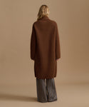A woman shown from the back wearing a long brown coat over wide-leg striped trousers, standing against a plain beige studio background.