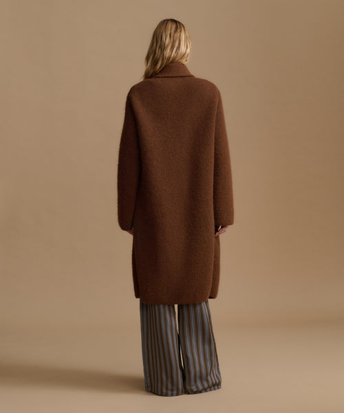 A woman shown from the back wearing a long brown coat over wide-leg striped trousers, standing against a plain beige studio background.