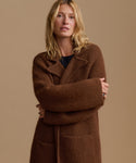 A woman wearing a long, oversized brown wool coat with wide lapels and a tie belt, posed against a solid beige studio background.